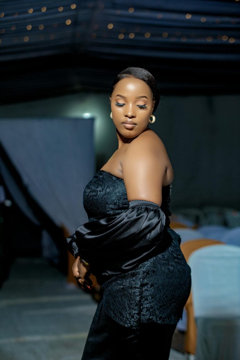 A stylish woman in a black dress poses elegantly at a nighttime indoor event.